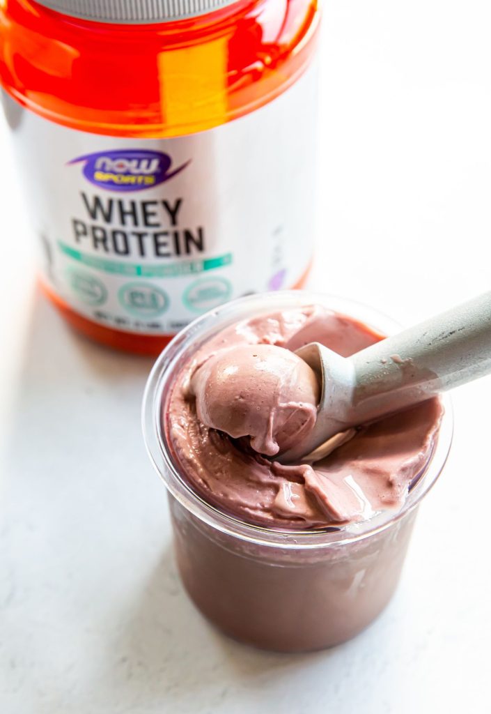 chocolate cherry ice cream in a clear pint with an ice cream scoop with a protein container in the background.